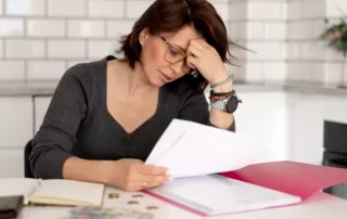 Woman wearing glasses sits at a kitchen table, stressed while reviewing documents, surrounded by coins, papers, and a pink folder — evoking anxiety about which assets she can keep in Chapter 13 bankruptcy.