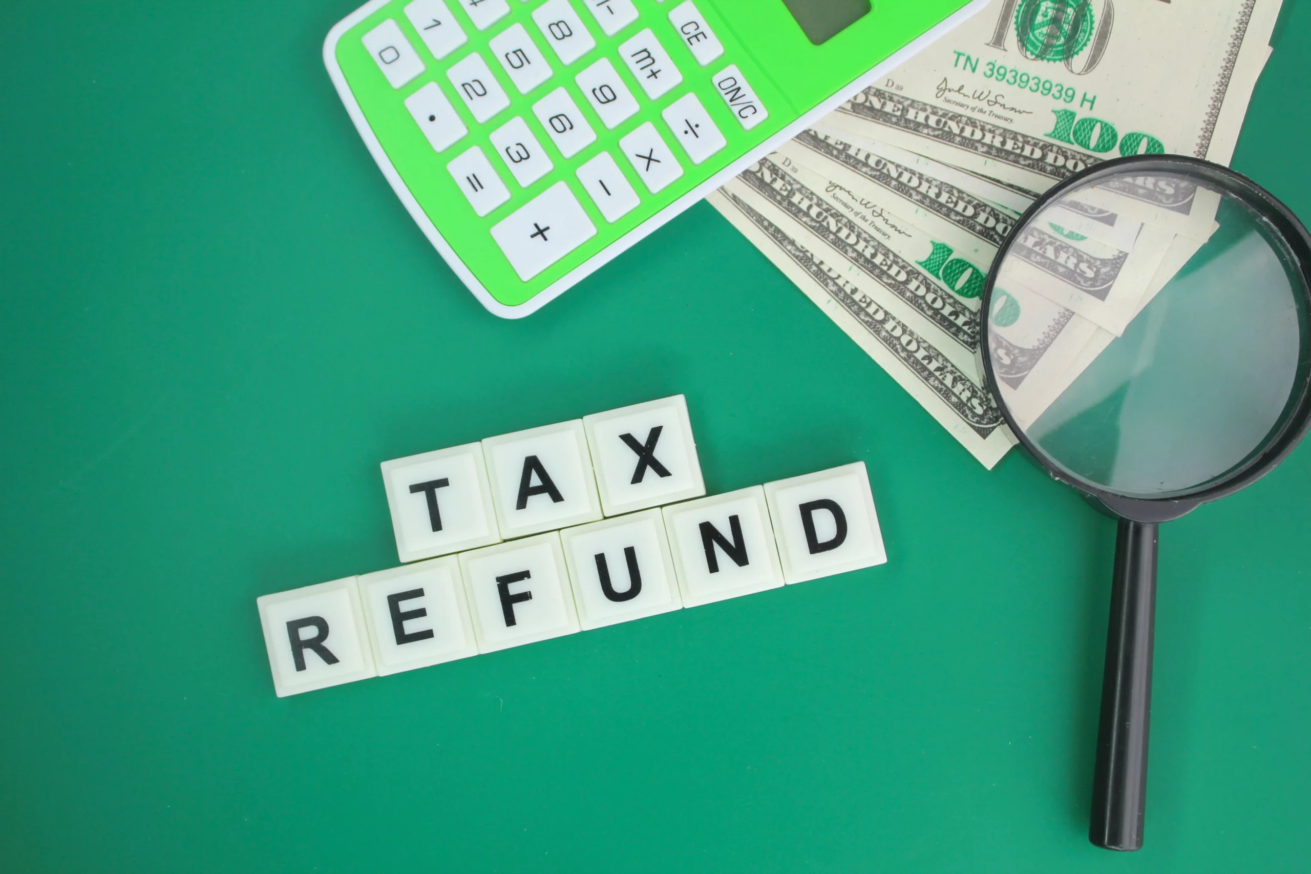 Calculator, magnifying glass, hundred‑dollar bills and Scrabble tiles spelling ‘TAX REFUND’ on green background, illustrating using a tax refund for Chapter 13 plan payments in New Jersey