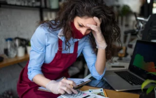 Worried woman in red apron and mask using calculator at cluttered desk with laptop graph, illustrating small business financial stress deciding between Chapter 7 and Chapter 11 in New Jersey.