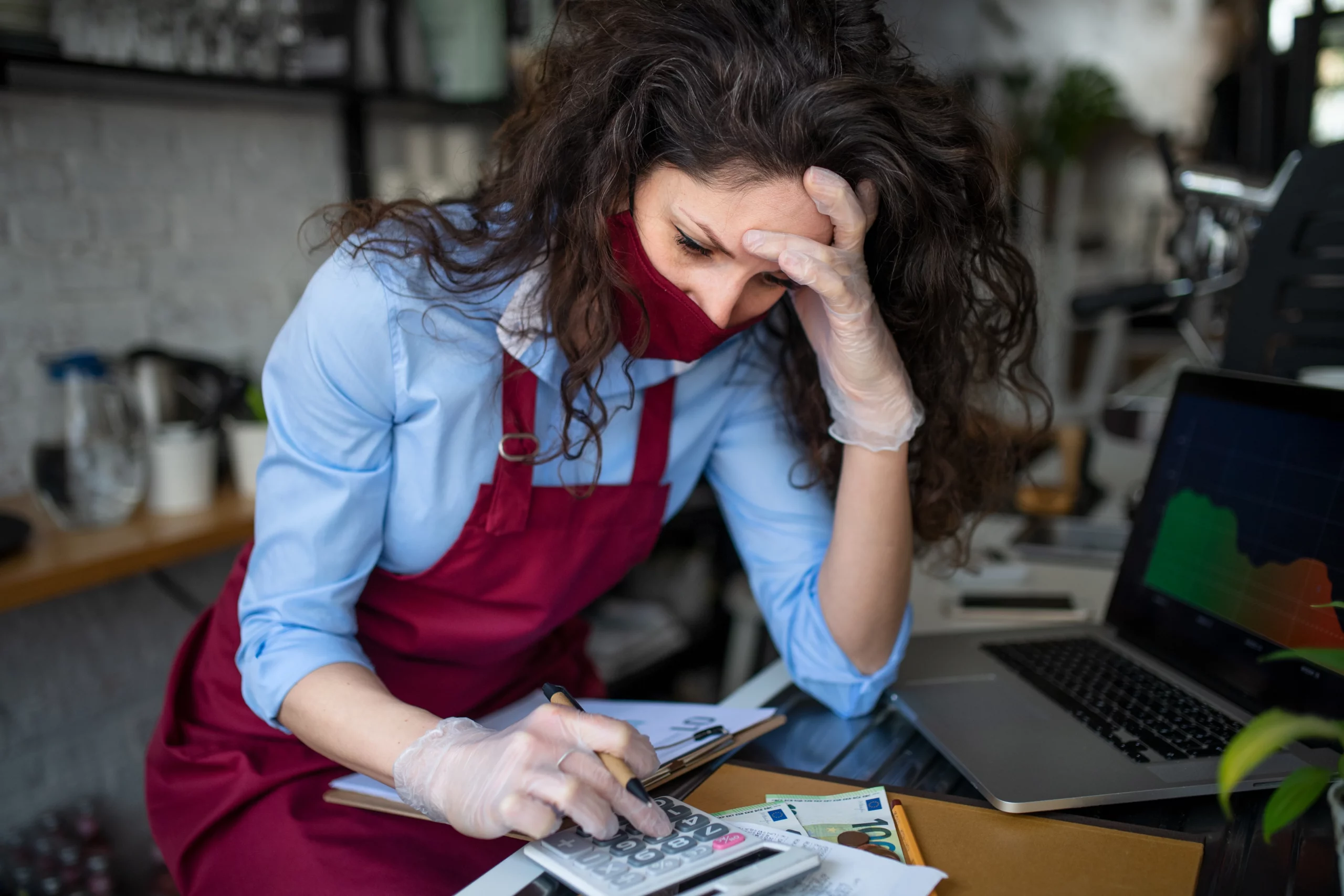 Worried woman in red apron and mask using calculator at cluttered desk with laptop graph, illustrating small business financial stress deciding between Chapter 7 and Chapter 11 in New Jersey.